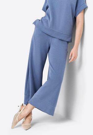 Persona con pantalones sueltos azules y top a juego apoyada contra una pared, con tacones slingback beige puntiagudos cruzados en los tobillos.