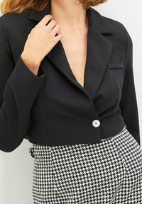 Przycięty czarny żakiet z wyłogami, jednolitym zapięciem na guzik i kieszenią naszytą. Połączony z spodniami we wzór houndstooth.