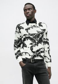 Schwarz-weiß gemusterter Strickpullover mit Rundhalsausschnitt, getragen über einem schwarzen Hemd mit Kragen, kombiniert mit dunklen Jeans und einem schwarzen Gürtel.