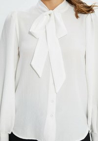 Femme portant un chemisier blanc à manches longues avec un grand nœud noué au cou, boutonné sur le devant.