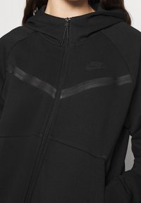 Woman wearing a black Nike zip-up hoodie with a subtle reflective stripe across the chest and a hood.