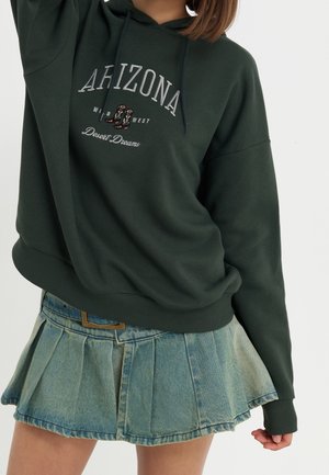 Donkergroene hoodie met "Arizona Wild West Desert Dreams" en cowboylaarzenafbeelding, gecombineerd met een lichtblauwe geplooide denim minirok.