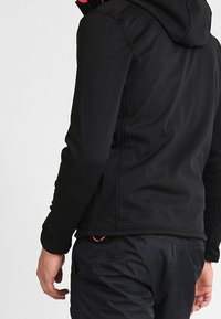 Chaqueta de softshell negra con capucha, que presenta una textura suave, bolsillos con cremallera y acentos rojos en el interior y en los cordones.