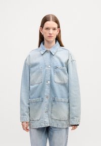 Veste en denim bleu clair avec fermeture à boutons, deux grandes poches avant et un col classique. Texture douce, coupe oversize.