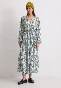 Blumenmuster Langarmkleid in Hellblau mit schwarzen und gelben Mustern, V-Ausschnitt, geraffte Taille und gestuftem Rock. Kombiniert mit schwarzen Loafers.