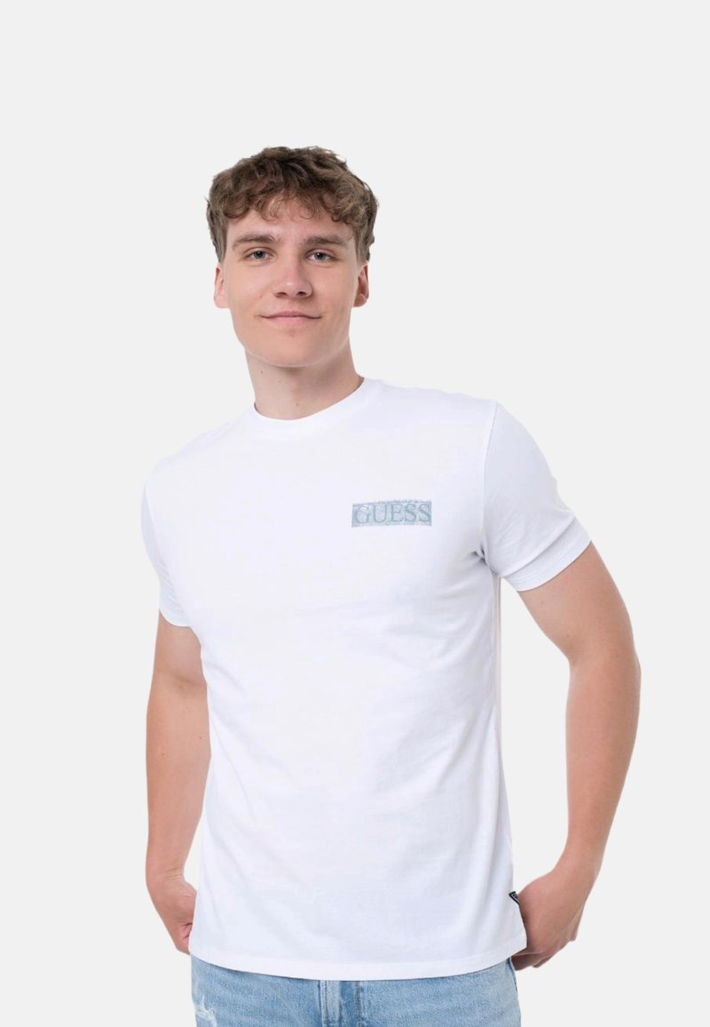 Camiseta de algodón blanca con un logo bordado en azul claro que dice "GUESS" en el lado izquierdo del pecho. Diseño de mangas cortas y cuello redondo.