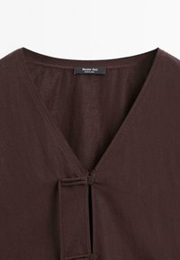 Gros plan sur un vêtement marron à col en V avec un détail de boutons à l'avant et une étiquette de marque visible indiquant Massimo Dutti, fabriqué en Chine.