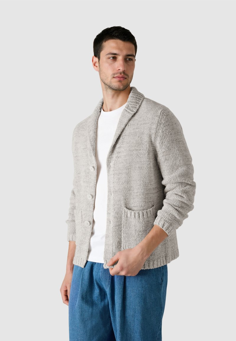 Mand iført lys gråstrikket cardigan over hvid skjorte og blå løse bukser, der kigger til siden mod en ensfarvet baggrund.