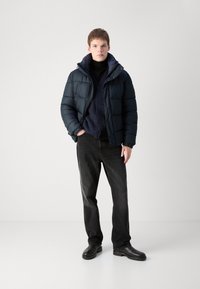 Doudoune marine avec texture matelassée, détails de boutons, superposée sur un pull en maille sombre, associée à un jean noir et des chaussures.