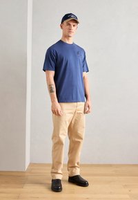 T-paita tummansinisestä puuvillasta, lyhyillä hihoilla ja pienellä logolla rinnassa, yhdistettynä vaaleanbeigeihin chinopö pantaloon ja mustiin liukkuihin kengiksi.