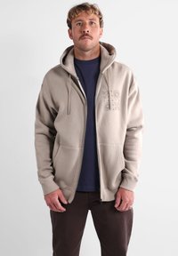 Haut de sweatshirt zippé beige avec une grande capuche, des poches avant et des motifs floraux sur le côté gauche de la poitrine, porté par-dessus un t-shirt bleu marine. Pantalons marron.