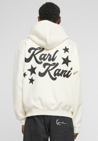 Sudadera crema con texto negro "Karl Kani" y gráficos de estrellas en la parte trasera. Cuenta con un bolsillo frontal y una capucha amplia.