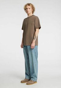 Rjava kratka majica s kratkimi rokavi in okrolim izrezom, kombinirana s svetlo modrimi širokimi kavbojkami in bež čevlji. Na rokah so vidne tetovaže.