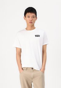 Tricou alb din bumbac cu mâneci scurte, guler rotund și patch cu logo-ul Levi's negru pe partea stângă a pieptului. Purtați cu pantaloni beji.