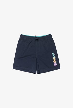 Niño con pantalones cortos de baño azul marino con cintura elástica y logotipo morado en la pierna izquierda, de pie descalzo contra un fondo blanco.