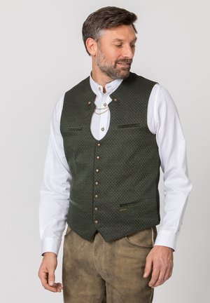 Man wearing dark green patterned vest with metal buttons and chain over white shirt and brown suede pants, looking to his right.