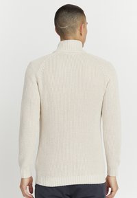 Beige strikkegenser med høy krage og ribbede mansjetter. Har et teksturert mønster og en avslappet passform. Bakvisningen viser sømløs skulderdesign.