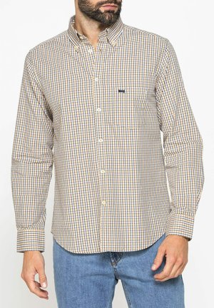 Uomo con indosso una camicia a maniche lunghe a quadri beige, blu e bianca, con jeans, mani rilassate ai lati, su sfondo semplice.