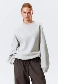 Sudadera gris de cuello redondo con mangas ragl�án, textura suave y puños ribeteados, combinada con pantalones oscuros de tiro con cordón marrón.