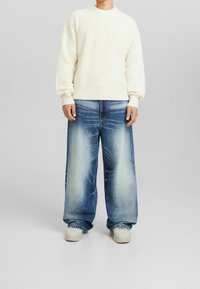 Osoba ubrana w biały sweter robiony na drutach, wyblakłe, luźne niebieskie dżinsy i białe sneakersy stoi na tle jednolitego szarego tła.