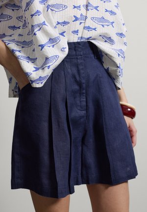 Persona que lleva pantalones cortos plisados de color azul marino con bolsillos laterales y una camisa blanca con estampado de peces azules, complementada con pulseras rojas y crema.