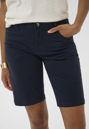 Sieviete, tērpusies tumši zilās ceļgalu garuma šortos un baltā, teksturētā topā, ar labo roku kabatā un zelta aproci uz kreisās plaukstas.