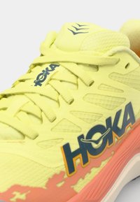 Baskets de sport jaune néon avec empeigne en maille, marque HOKA bien en vue, et un dégradé rose-orange sur la semelle. Design texturé et léger.