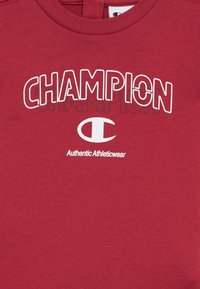 Κόκκινο βαμβακερό φούτερ με έντονα λευκά γράμματα "CHAMPION" και κυκλικό λογότυπο.  Μαλακή υφή με στρογγυλή λαιμόκοψη και κοντά μανίκια.