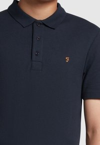 Marineblaues Polohemd aus geripptem Stoff mit klassischem Kragen, drei Knöpfen und einem orangefarbenen Logo auf der linken Brust.