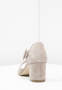 Scarpa alta in suede beige chiaro, con punta arrotondata, cinturino regolabile e un distintivo dettaglio a bottone sul lato esterno del tacco.