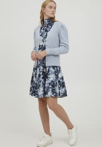 Cardigan en maille côtelée gris clair avec boutons, porté par-dessus une robe fleurie bleu marine. Des baskets blanches complètent la tenue, prise en pose debout devant un fond uni.