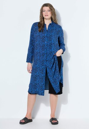 Kobieta stoi przy białej ścianie, ubrana w niebieską długą sukienkę koszulową zapinaną na guziki z czarnymi cętkami lamparta, czarne szorty i czarne klapki na płaskiej podeszwie.