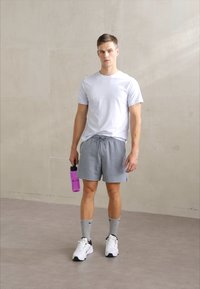 Ținută sportivă formată dintr-șoie gri, tricou alb cu mânecă scurtă, șosete gri și adidași negru-alb, ținând o sticlă de apă roz.