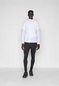 Giacca leggera bianca con chiusura a zip, leggings neri aderenti e scarpe da ginnastica nere. Indossata con occhiali da sole blu a specchio.