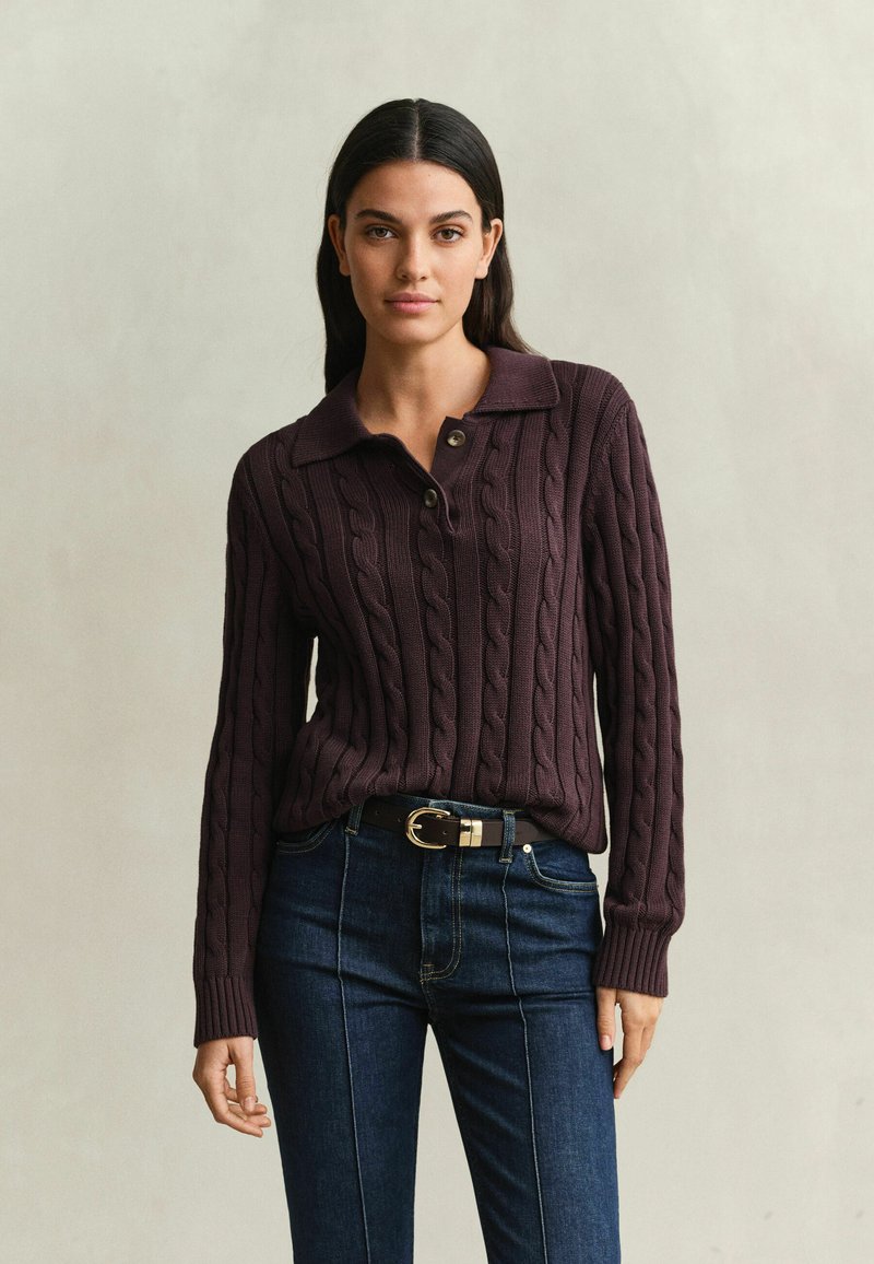 Femme aux longs cheveux foncés portant un pull torsadé violet foncé avec col et un jean bleu foncé avec une ceinture noire, debout devant un fond uni.
