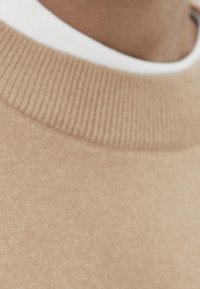 Beige tröja med ribbad halsringning och mjuk textur, med ett vitt innerlager synligt vid kragen som framhäver färgkontrasten.