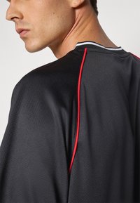 T-shirt de sport noir avec une finition texturée, des rayures d'accent rouge et un col côtelé avec des détails blancs, conçu pour un usage athlétique.