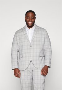 Traje de cuadros grises con chaqueta de un botón, dos bolsillos frontales y pantalones a juego. Camisa de vestir blanca debajo. Textura suave.