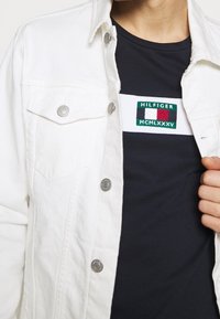 Bílá džínová bunda se stříbrnými knoflíky, nošená přes námořnické tričko s zelenou, bílou a červenou nášivkou s textem.