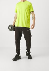 Neonově zelené sportovní tričko s krátkým rukávem; černé tepláky s logem; drží činku; má na sobě černé sportovní boty. Jednoduchý, sportovní design.