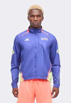 Mann mit kurzen blonden Haaren trägt eine violette BMW Berlin Marathon Jacke und leuchtend orange Shorts und steht vor einem einfarbigen Hintergrund.