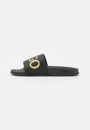 Black slide sandals featuring a wide strap with a gold logo, textured insole, and rubber outsole with a ridged pattern for grip.