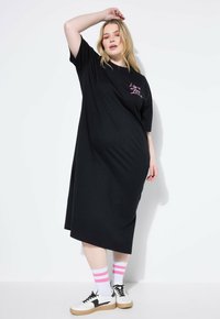 Czarna, oversize sukienka t-shirt z krótkimi rękawami, z różowym napisem. Połączona z białymi trampkami i paskowanymi różowo-szarymi skarpetkami.