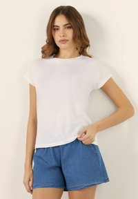 Tricou alb cu mânecă scurtă dintr-un material ușor, având un guler rotund și o croială lejeră, asortat cu pantaloni scurți din denim albastru.