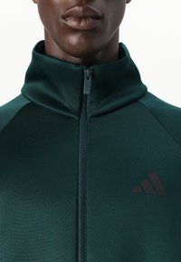 Sötétzöld, zippes dzseki magas gallérral, sima anyagból. Az öltönyön fekete Adidas logó található a mellrészen és tónusos varrás részletezéssel rendelkezik.