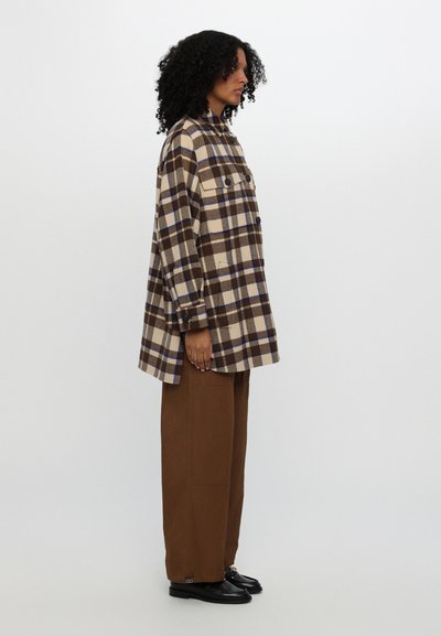 Chaqueta de camisa a cuadros marrón y beige con dos bolsillos frontales, detalles de botones y corte holgado, combinada con pantalones anchos marrones y zapatos negros.
