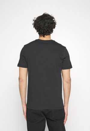 Homme aux cheveux bouclés et foncés portant un t-shirt noir uni à manches courtes et un pantalon noir, debout face à un fond blanc.