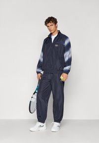 Men's navy tracksuit featuring a zip jacket with blue gradient sleeves and elasticated cuffs, paired with matching trousers and white sneakers.