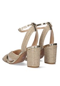 Sandalias de tacón alto en dorado y beige con parte superior de tela texturizada, detalles metálicos y un tacón grueso decorado con dorados. Diseño de punta abierta.