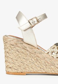 Sandale compensée avec une plateforme en jute tressé, une sangle en cuir métallique doré, et des accents tissés décoratifs sur le dessus. Fermeture à boucle.